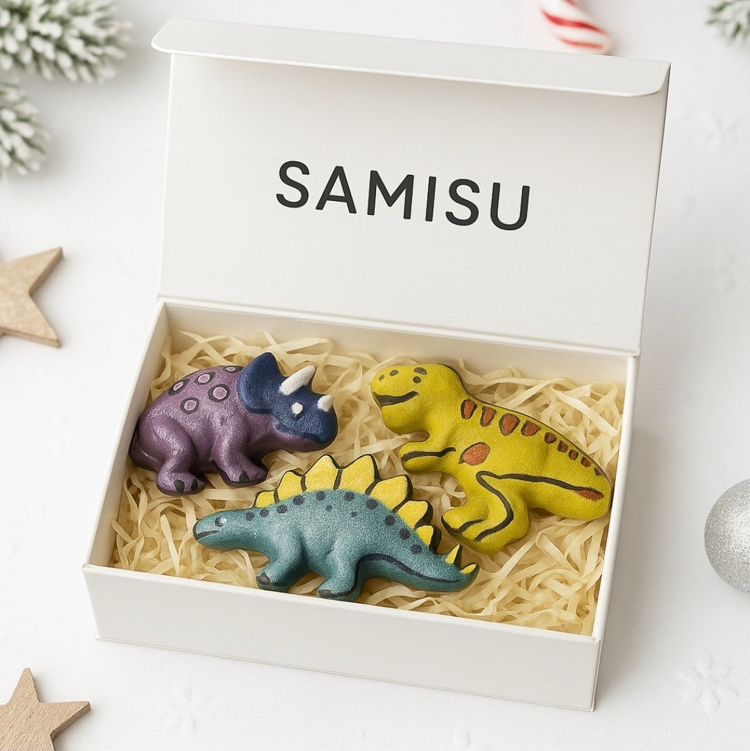 Three dinosaur-shaped bath bombs in a box labeled 'SAMISU' on a white surface.